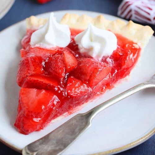 Slice of strawberry pie with jello topped with whipped cream on a plate with a fork on the side.