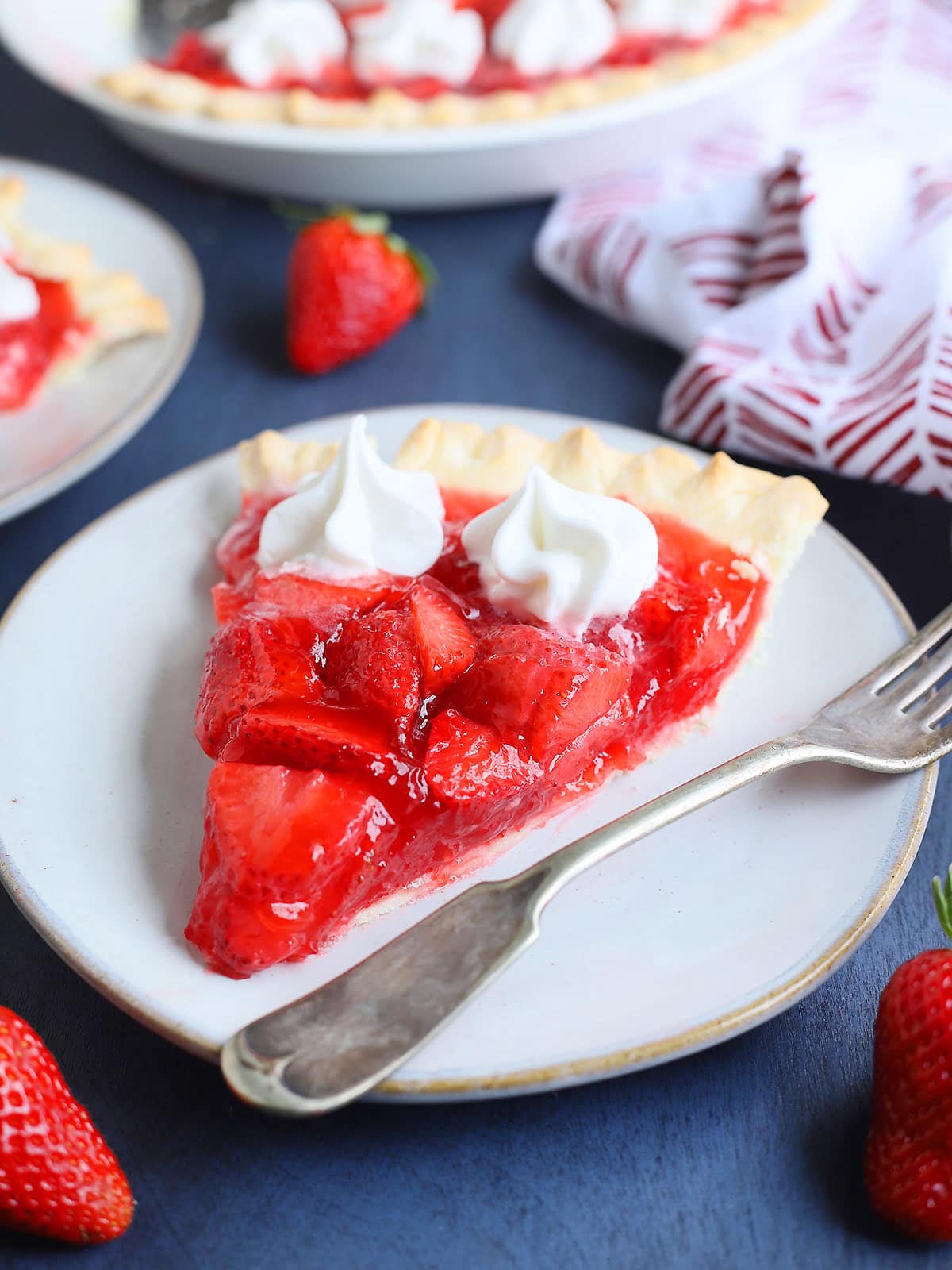 Slice of strawberry pie with jello topped with whipped cream on a plate with a fork on the side.