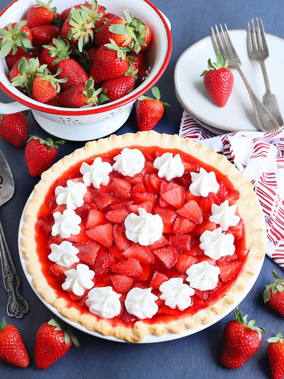 Whole strawberry pie with jello topped with whipped cream with a colander of fresh strawberries, plates, and forks on the side.