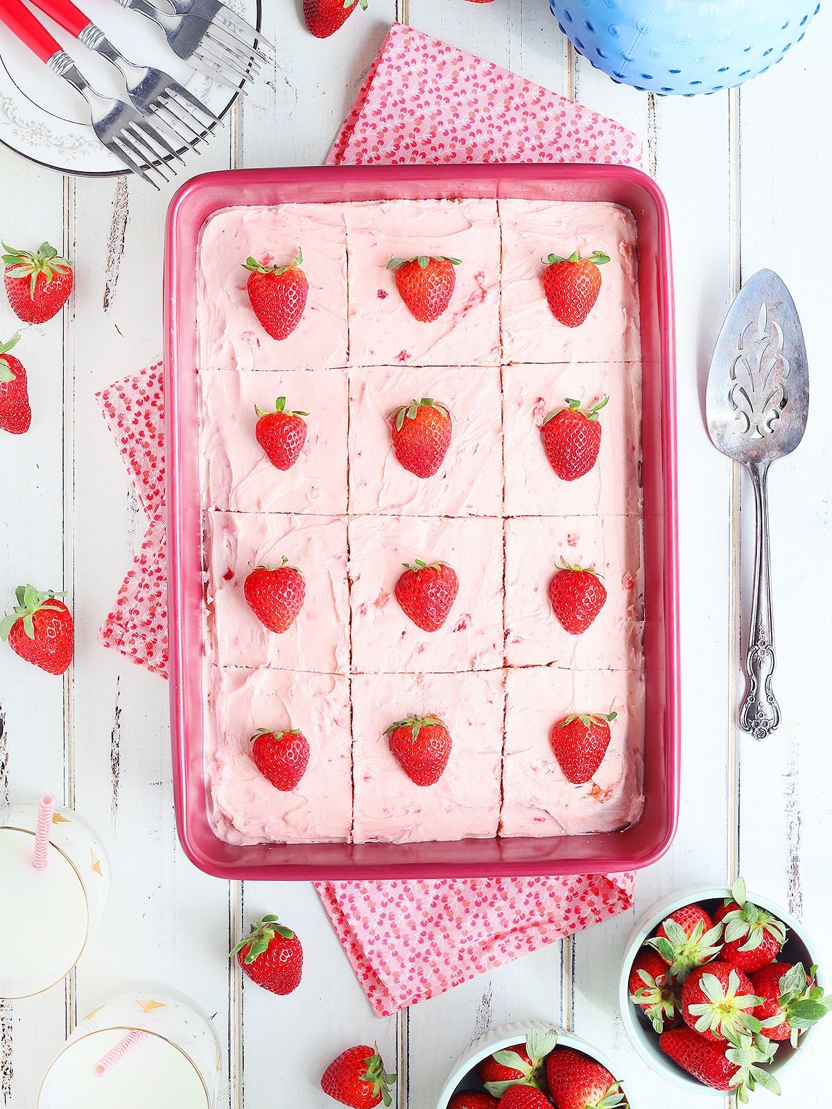 Cake pan of strawberry sheet cake with a dish of fresh strawberries, a cake server and glasses of milk on the side.