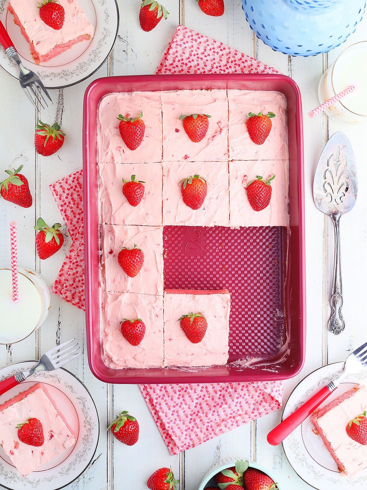 Cake pan of strawberry sheet cake with three slices missing. Slices of cake and glasses of milk to the side.