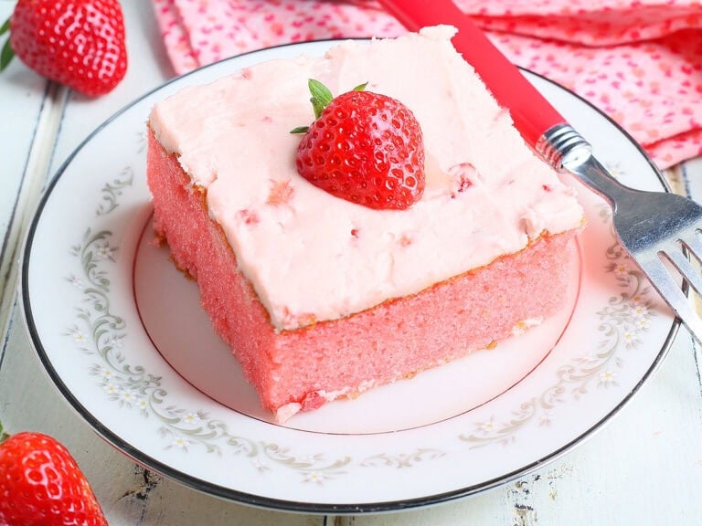 Single slice of strawberry sheet cake garnished with a fresh strawberry slice on a plate with a fork to the side.