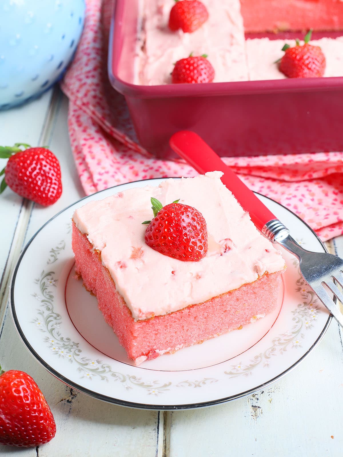 Single slice of strawberry sheet cake garnished with a fresh strawberry slice on a plate with a fork to the side.