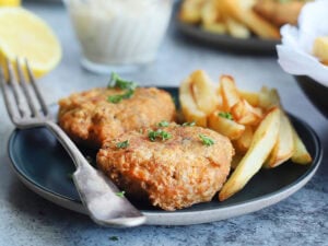 Two fried salmon croquettes garnished with fresh parsley on a plate with a fork and some French fries on the side. A dish of tartar sauce, a second plate of food and a platter of cooked salmon croquettes can be seen in the background.