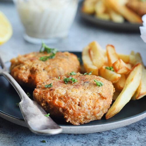 Two fried salmon croquettes garnished with fresh parsley on a plate with a fork and some French fries on the side. A dish of tartar sauce, a second plate of food and a platter of cooked salmon croquettes can be seen in the background.