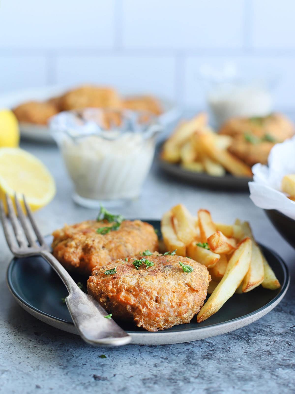 Two fried salmon croquettes garnished with fresh parsley on a plate with a fork and some French fries on the side. A dish of tartar sauce, a second plate of food and a platter of cooked salmon croquettes can be seen in the background.