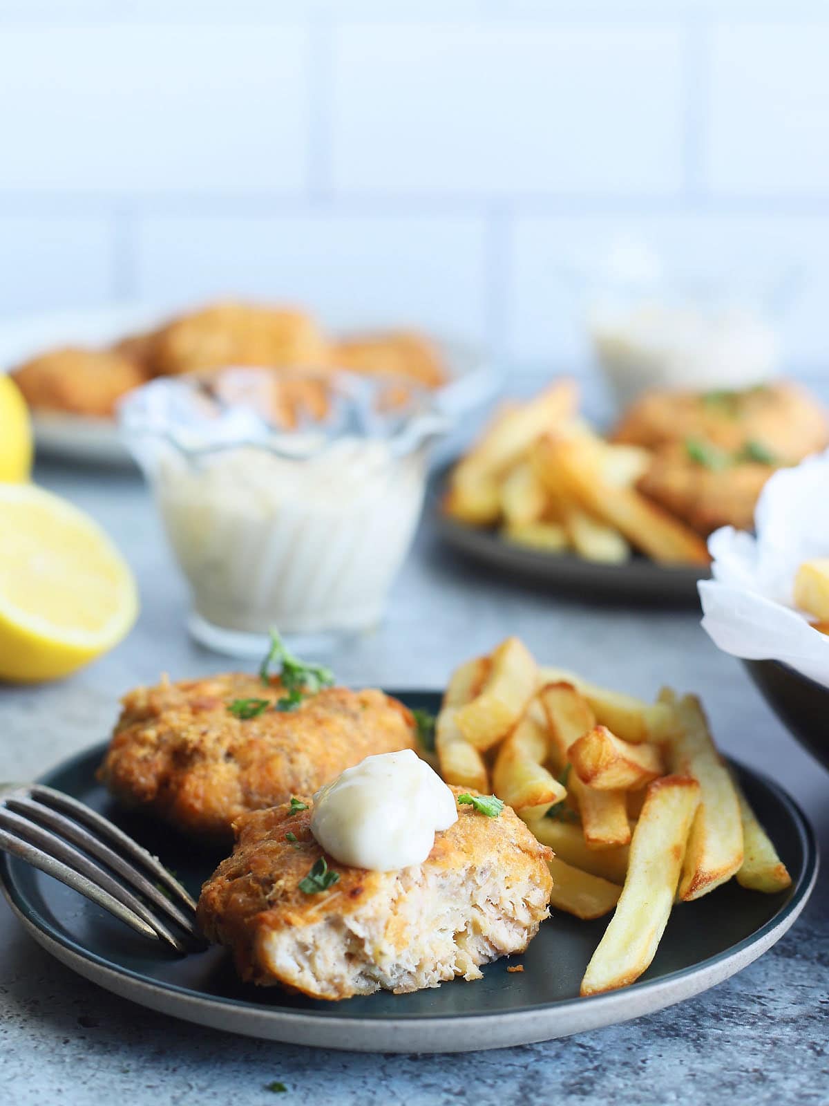 Two fried salmon croquettes garnished with fresh parsley on a plate with a fork and some French fries on the side. One of the croquettes has been cut in half and is garnished with a dollop of tartar sauce. A dish of tartar sauce, a second plate of food and a platter of cooked salmon croquettes can be seen in the background.