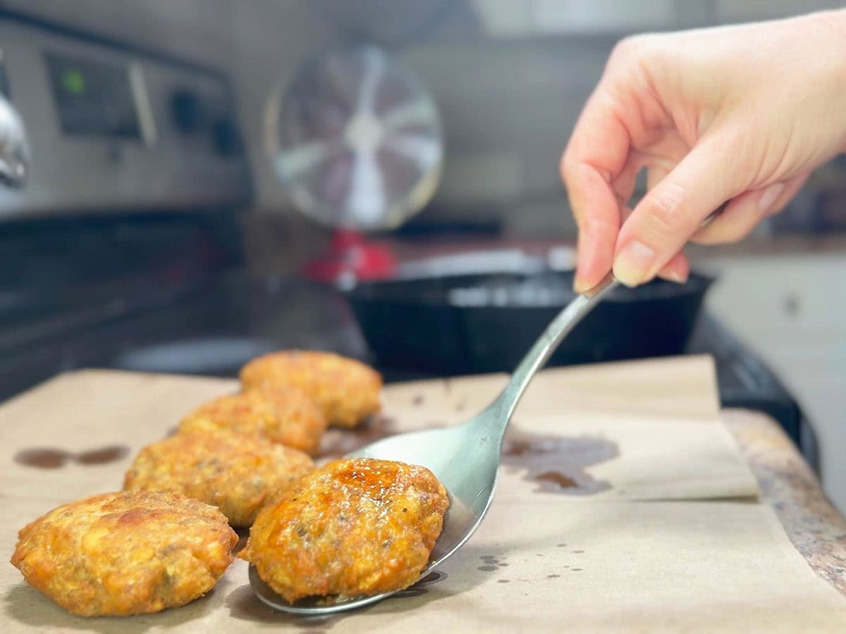 Hand transferring a fried salmon croquette from the skillet to a paper sack to drain.
