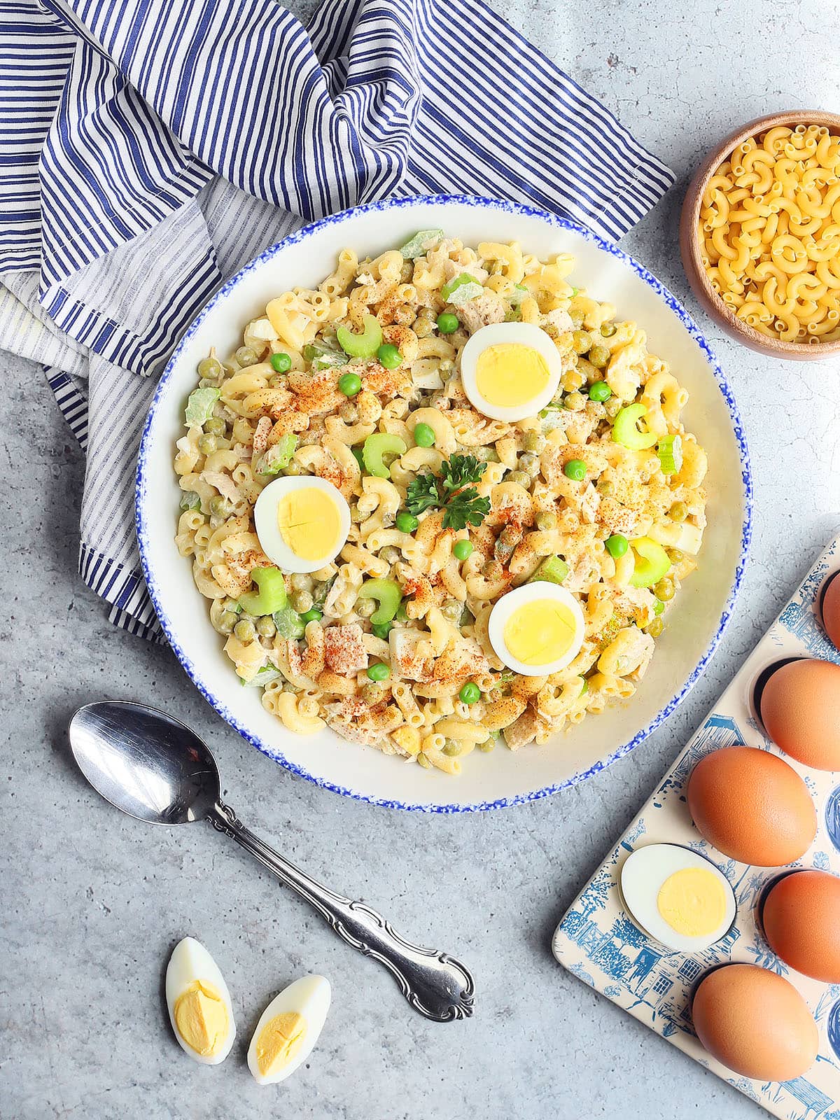 Serving bowl of tuna macaroni salad garnished with sliced hard boiled eggs, chopped celery, green peas, a sprig of parsley and a sprinkling of paprika. A small dish of macaroni pasta, sliced hard boiled eggs, a serving spoon and a napkin can be seen off to the side.