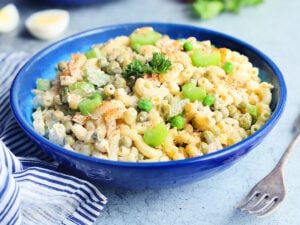 A close-up of a bowl of tuna macaroni salad. The salad contains elbow macaroni, green peas, chopped celery, and a creamy dressing, garnished with a sprig of parsley and a sprinkle of paprika. In the background, another bowl of salad, two brown eggs, celery stalks, and a halved boiled egg are visible with a striped cloth napkin nearby.