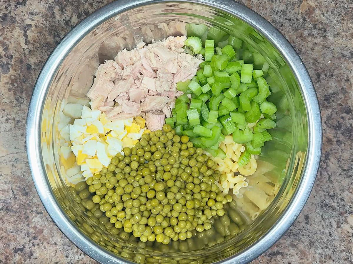 Cooked macaroni noodles, chopped hard boiled eggs, chunks of tuna, diced celery, and sweet peas in a large mixing bowl.