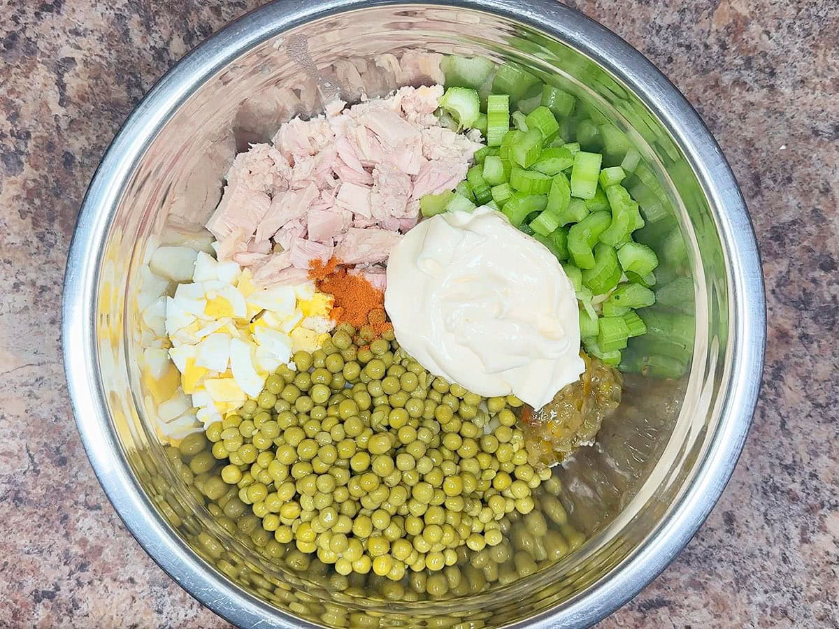 Cooked macaroni noodles, chopped hard boiled eggs, chunks of tuna, diced celery, sweet peas, pickle relish, cayenne pepper, and mayonnaise in a large mixing bowl.