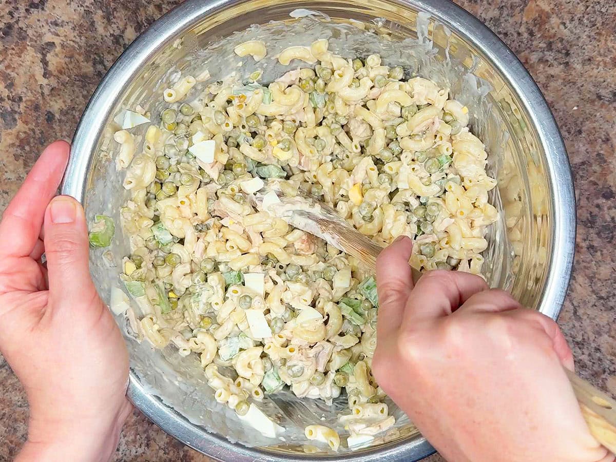 Hands using a wooden spoon to stir the tuna macaroni salad ingredient together.