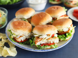 Platter of grilled catfish sliders with lettuce, sauce, tomatoes, slaw and potato chips in the background.