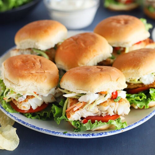 Platter of grilled catfish sliders with lettuce, sauce, tomatoes, slaw and potato chips in the background.