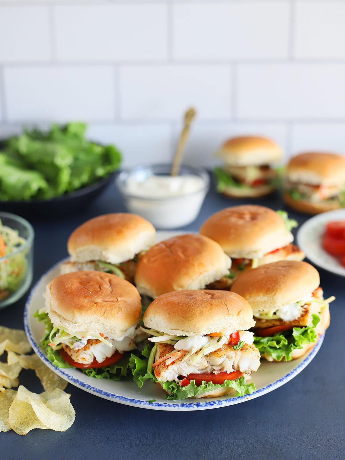 Platter of grilled catfish sliders with lettuce, sauce, tomatoes, slaw and potato chips in the background.