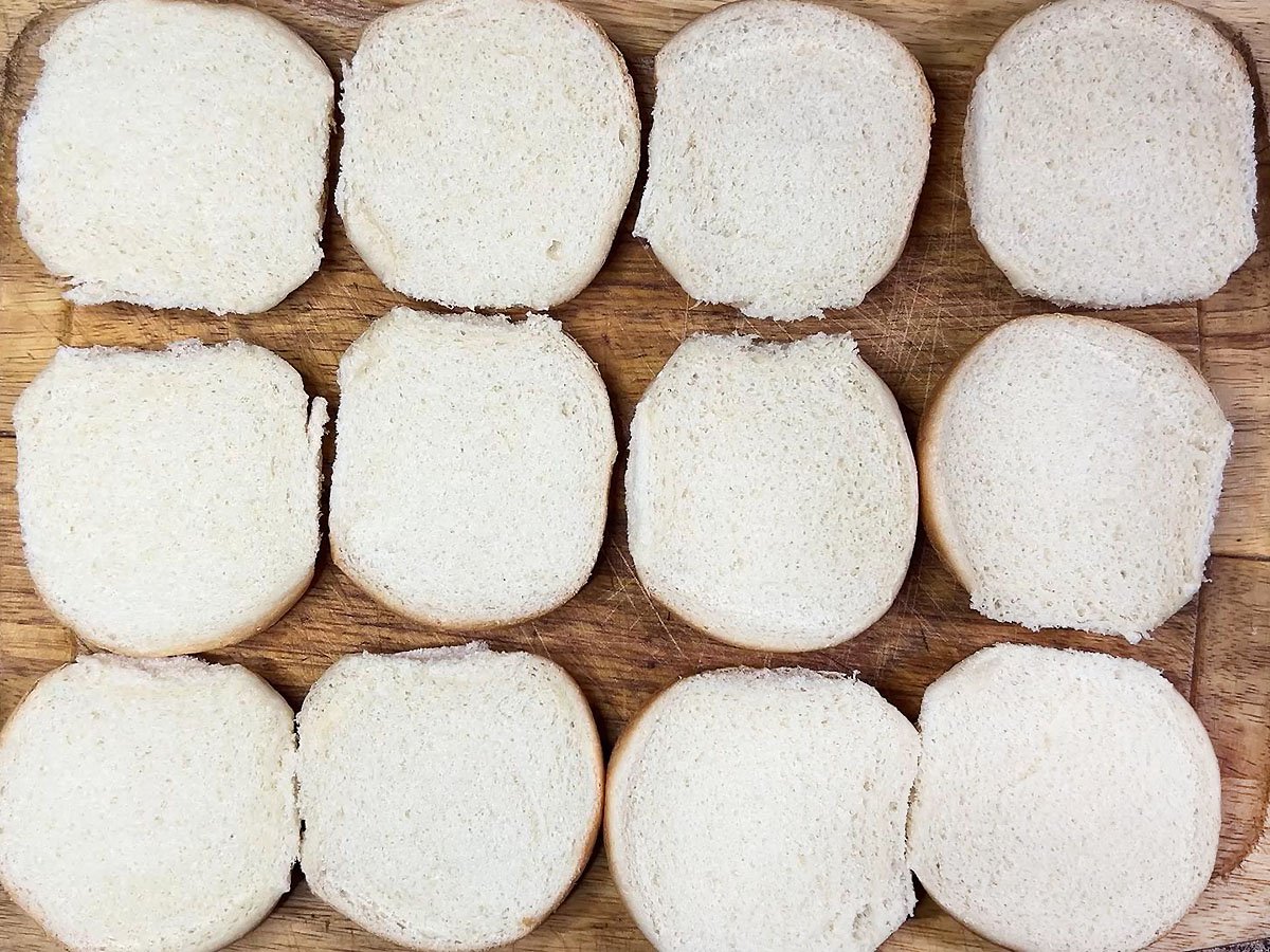 Bottom half of 12 slider buns on a wooden cutting board.