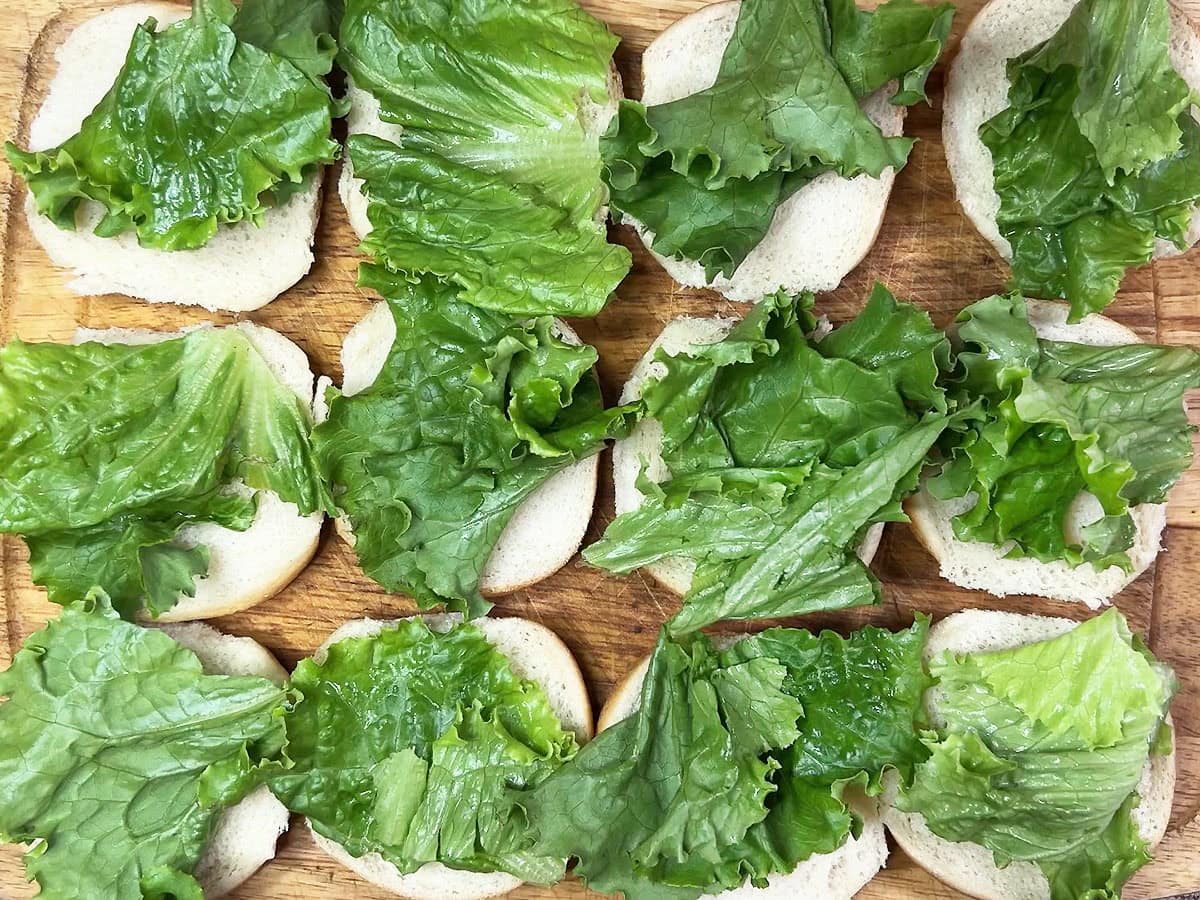 Bottom half of 12 slider buns topped with lettuce on a wooden cutting board.