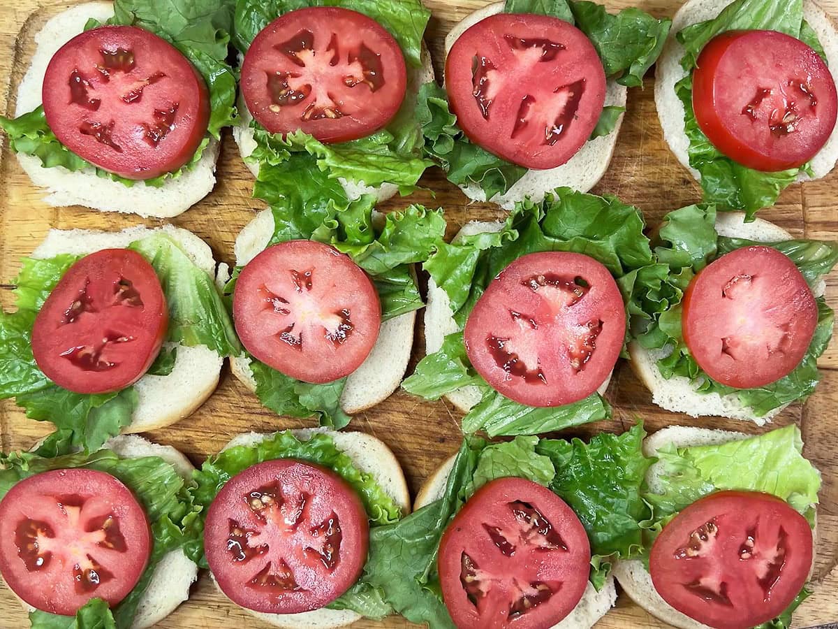 Bottom half of 12 slider buns topped with lettuce and tomato on a wooden cutting board.