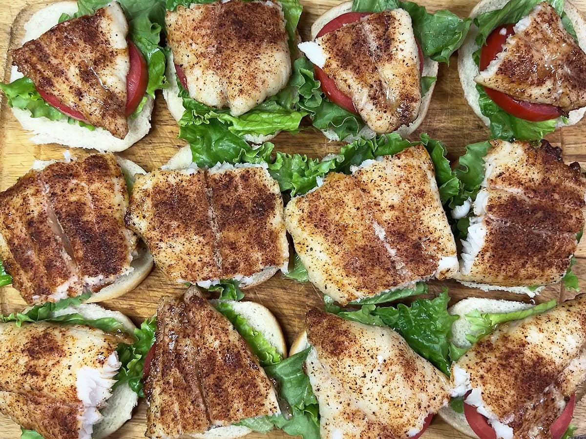 Bottom half of 12 slider buns topped with lettuce, tomato and a piece of grilled catfish on a wooden cutting board.