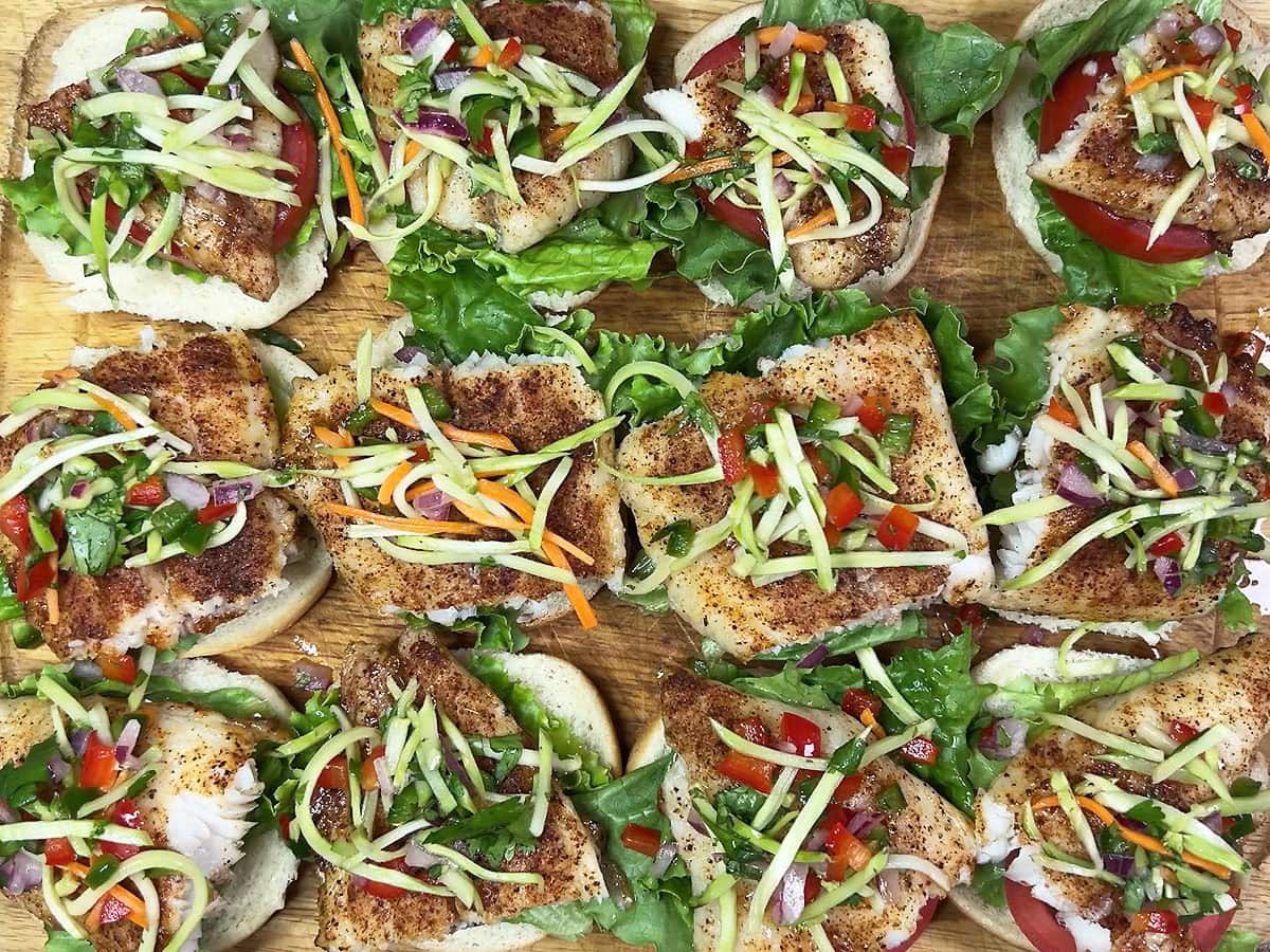 Bottom half of 12 slider buns topped with lettuce, tomato, a piece of grilled catfish, and coleslaw on a wooden cutting board.
