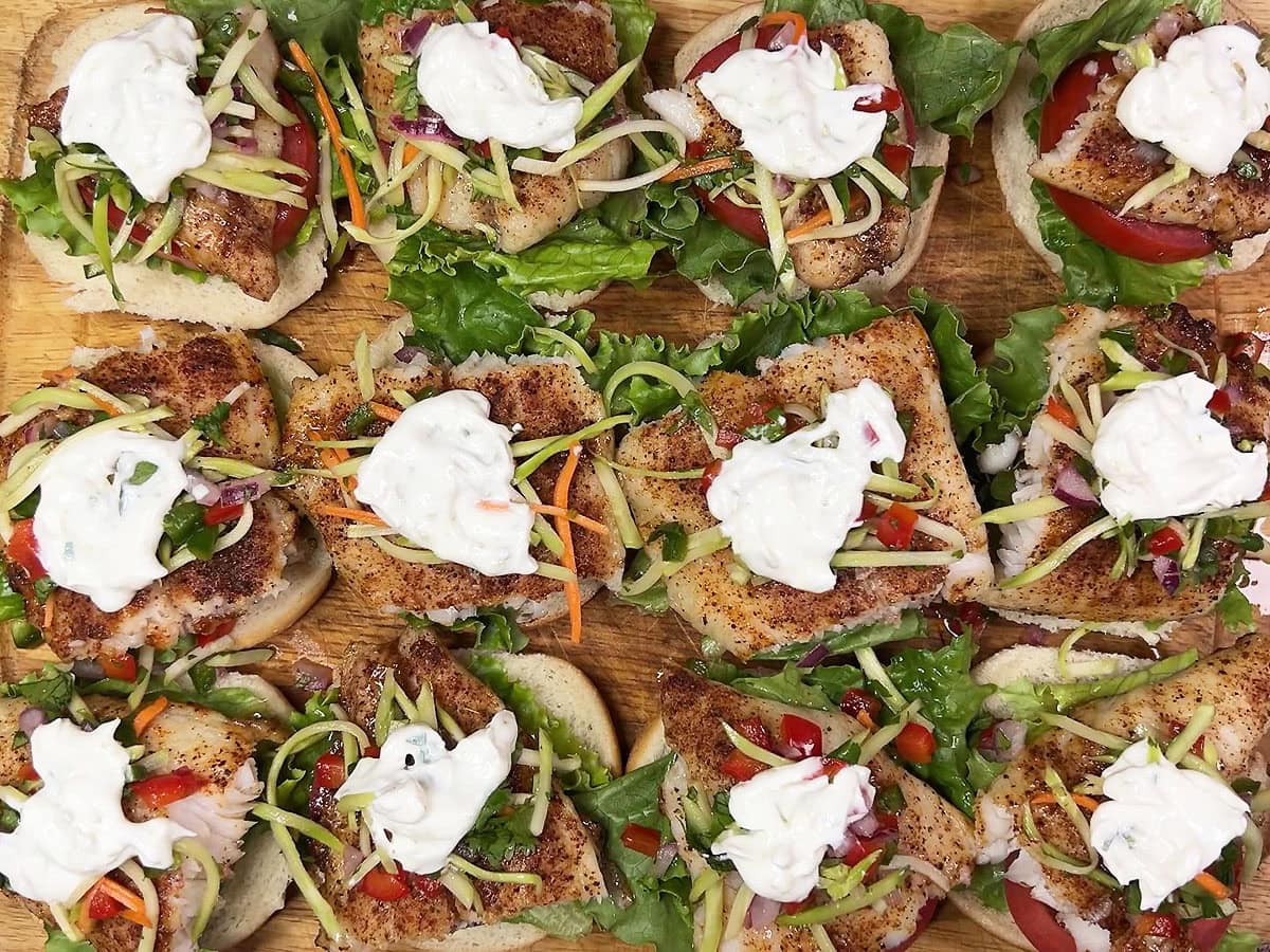 Bottom half of 12 slider buns topped with lettuce, tomato, a piece of grilled catfish, coleslaw, and creamy lime sauce on a wooden cutting board.