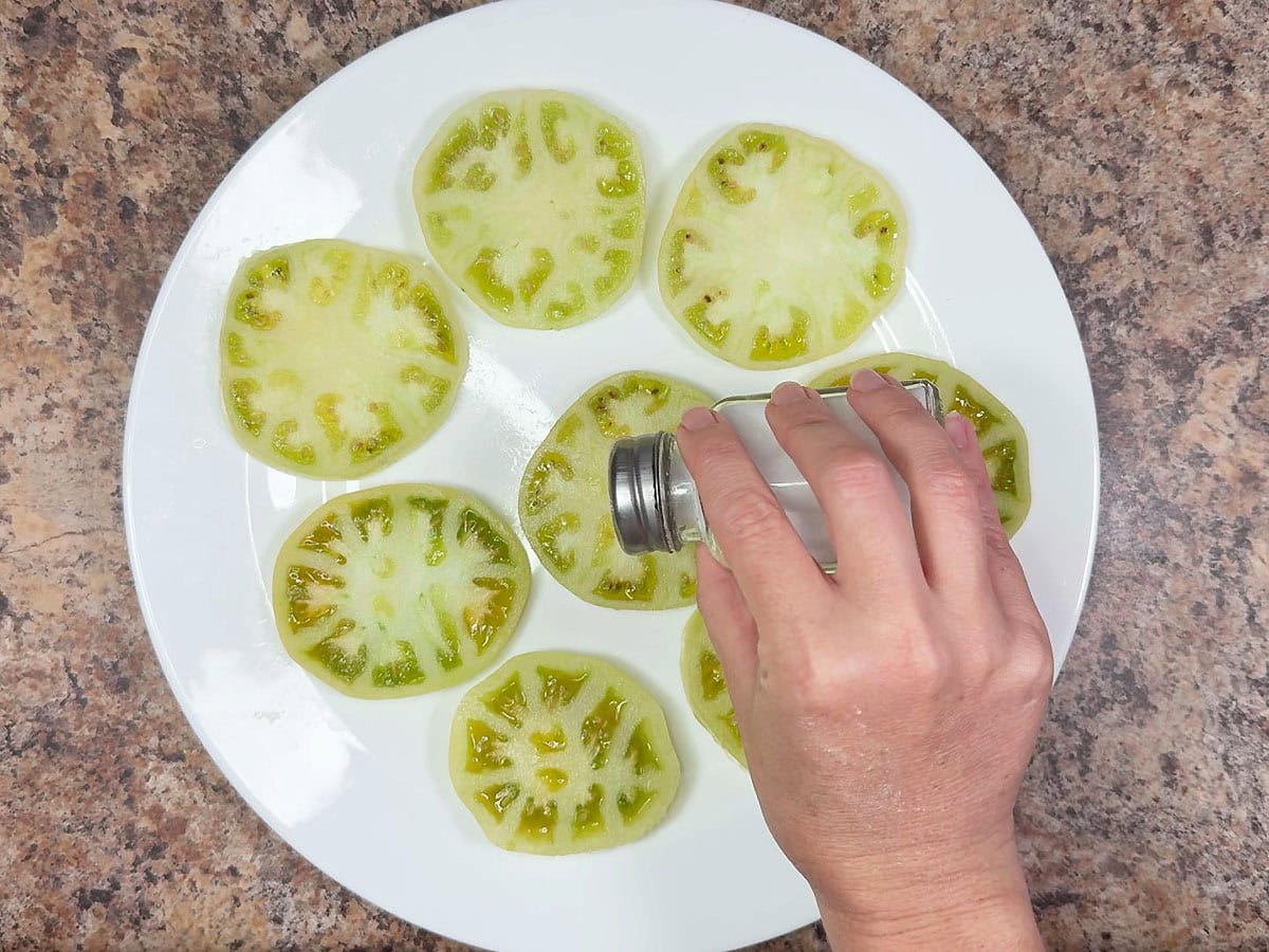 Sprinkling salt over a plate of sliced green tomatoes.
