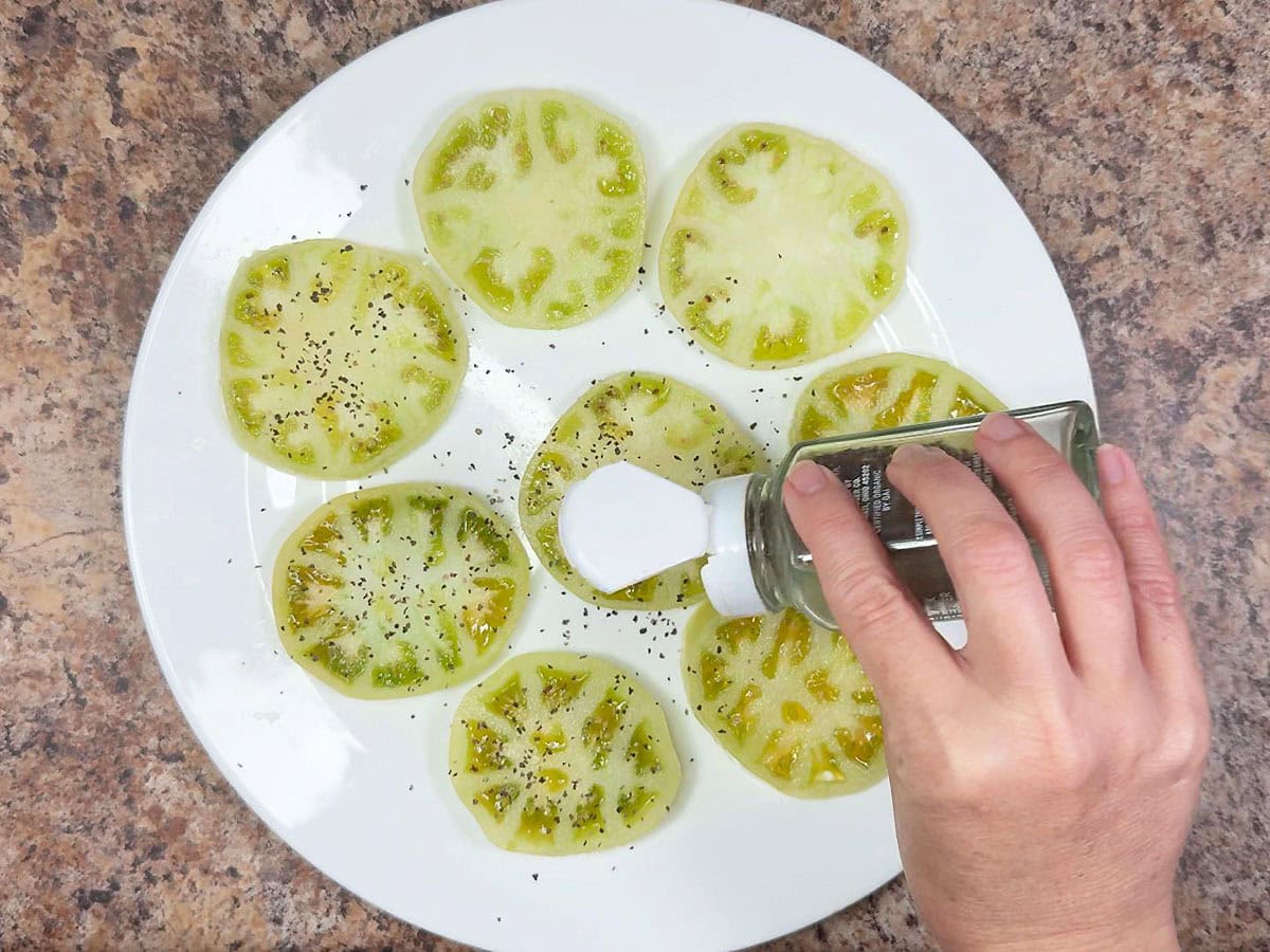 Seasoning the outside of sliced green tomatoes with ground black pepper.