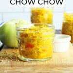Small jar of green tomato chow chow on a wooden cutting board with a green tomato and taller jars of chow chow in the background.