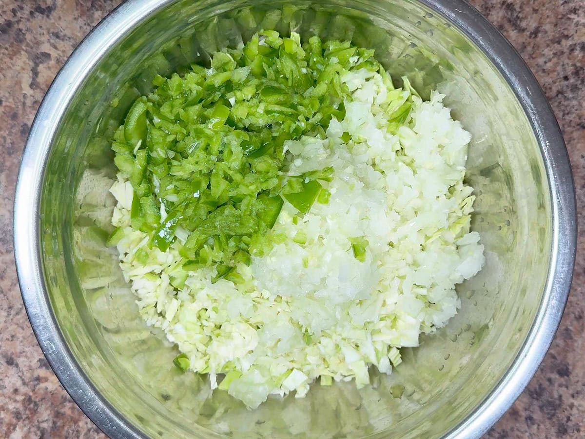 Diced green bell pepper added to the onions,shredded green cabbage, and diced green tomatoes in a metal mixing bowl.