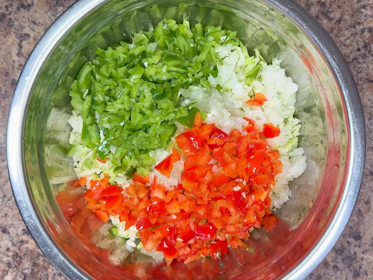 Diced red bell pepper added to the diced green bell pepper, onions,shredded green cabbage, and diced green tomatoes in a metal mixing bowl.