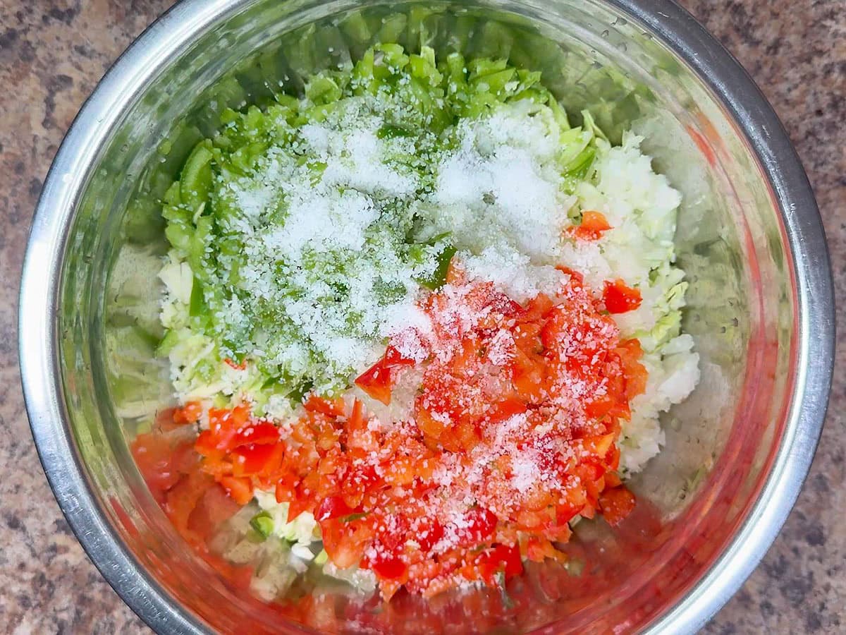 Kosher salt added to the diced bell peppers, onions,shredded green cabbage, and diced green tomatoes in a metal mixing bowl.