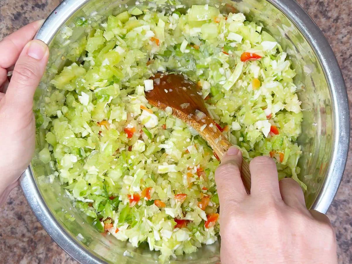 Stirring the cabbage, bell peppers, onions, green tomatoes, and kosher salt together.