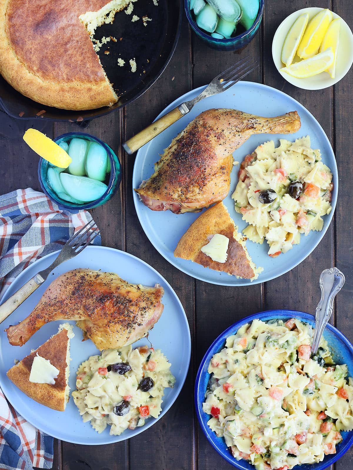 Two smoked chicken quarters on plates with Greek pasta salad and a wedge of cornbread topped with a pat of butter.