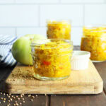 Small jar of green tomato chow chow on a wooden cutting board with a green tomato and taller jars of chow chow in the background.