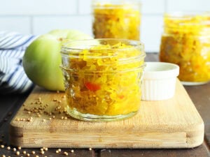Small jar of green tomato chow chow on a wooden cutting board with a green tomato and taller jars of chow chow in the background.
