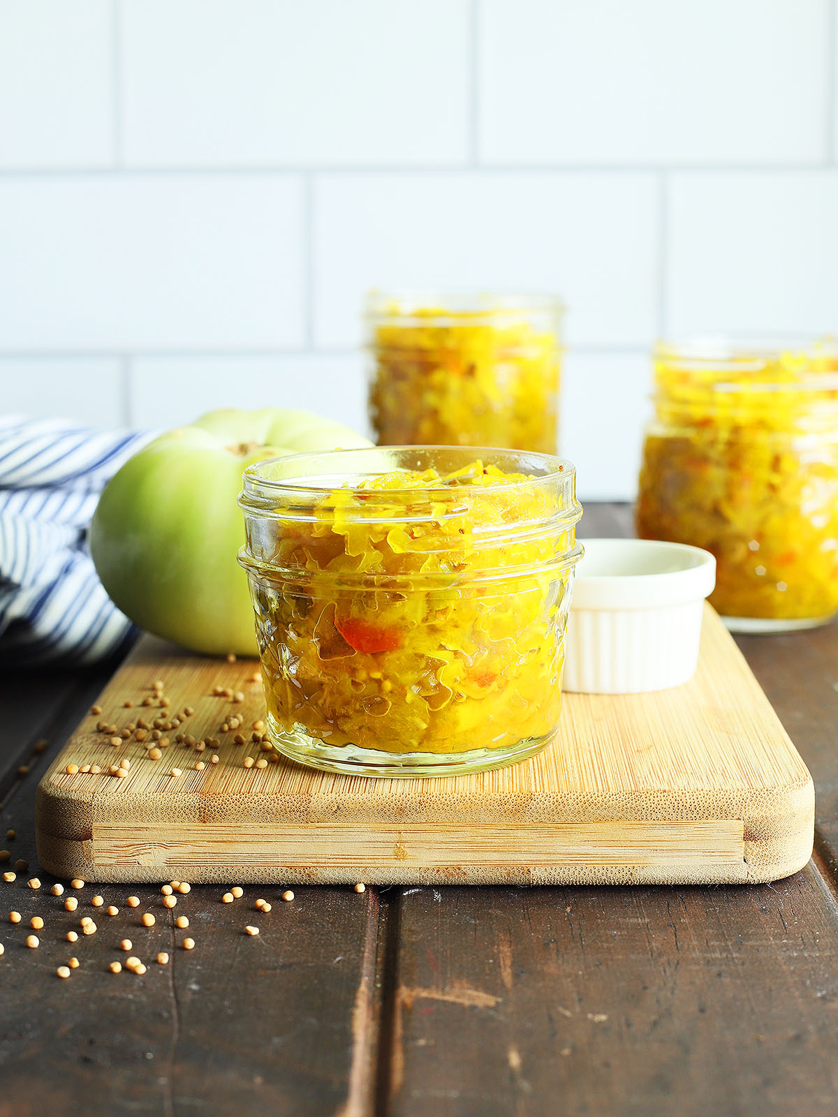 Small jar of green tomato chow chow on a wooden cutting board with a green tomato and taller jars of chow chow in the background.
