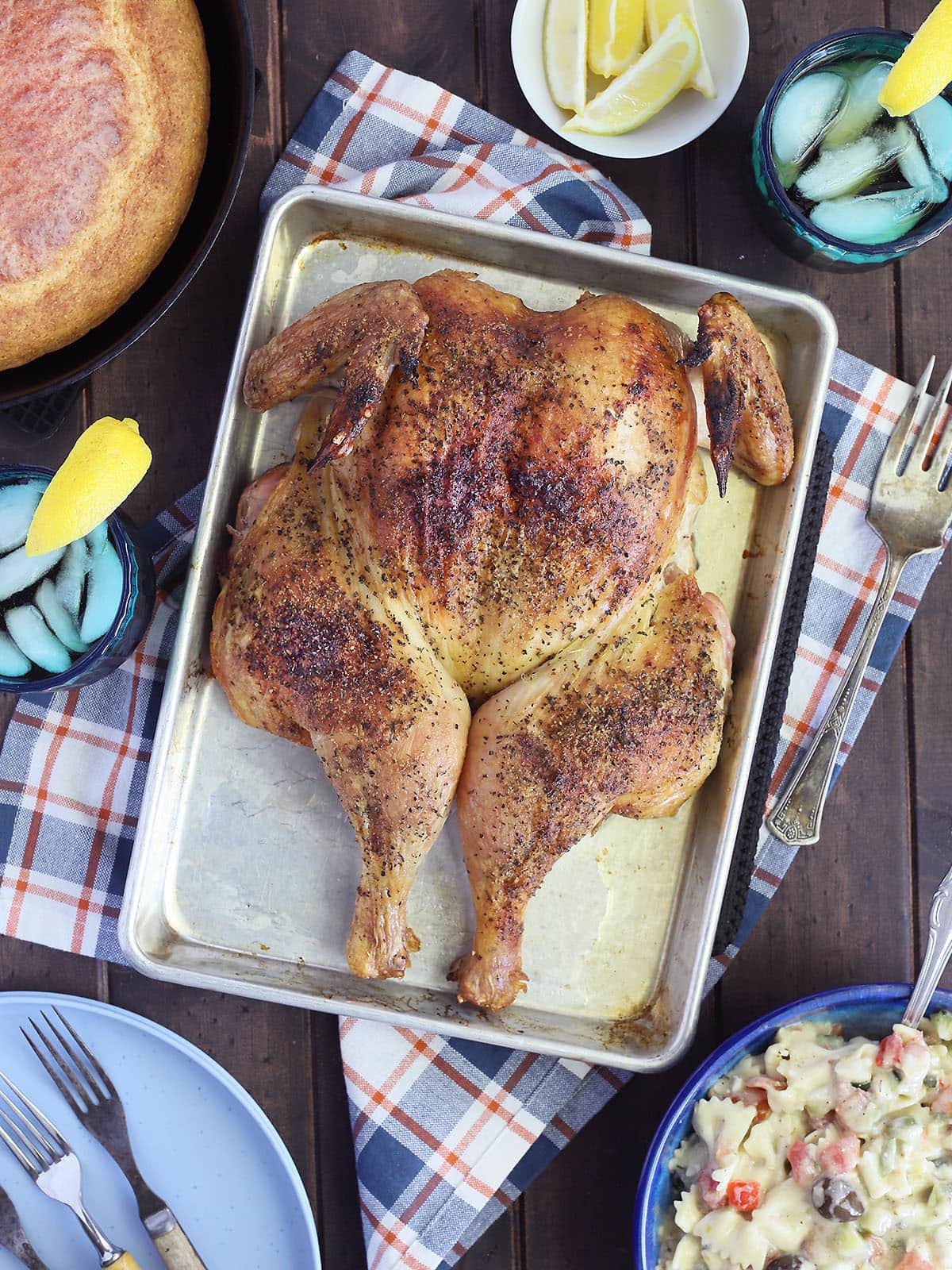 Smoked spatchcock chicken on a tray with a skillet of cornbread, a bowl of pasta salad, and glasses of sweet tea on the side