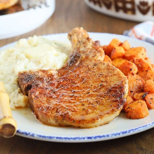 Seasoned air fryer bone in pork chop on a plate with mashed potatoes, roasted carrots and a fork on the side.