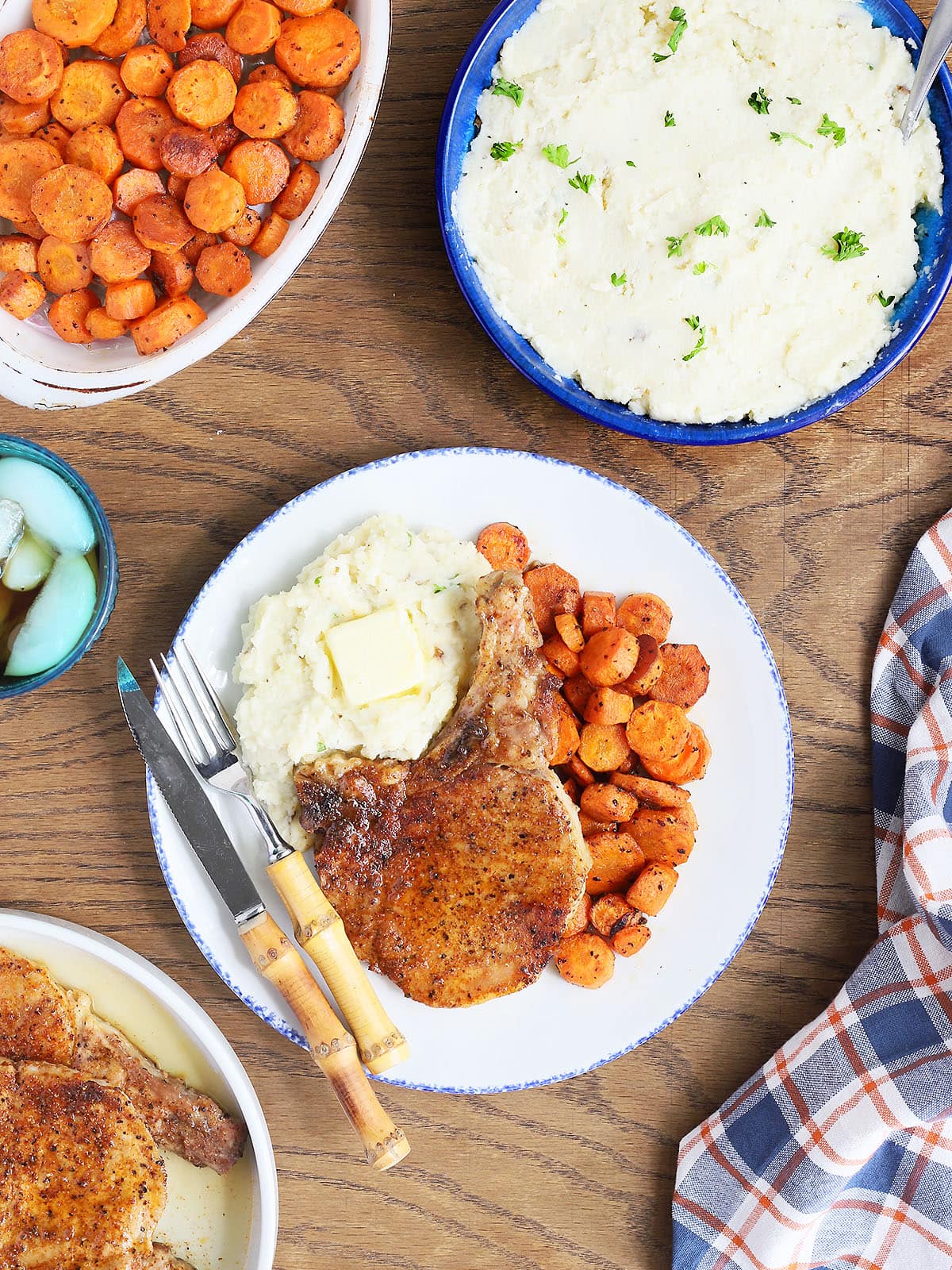 Air fryer bone in pork chop on a plate with mashed potatoes and roasted carrots with a knife and a fork positioned on the side. Serving dishes of potatoes, carrots and additional pork chops can be seen.
