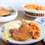 Air fryer bone in pork chop on a plate with mashed potatoes and roasted carrots with a knife and a fork positioned on the side. Serving dishes of carrots and additional pork chops can be seen.