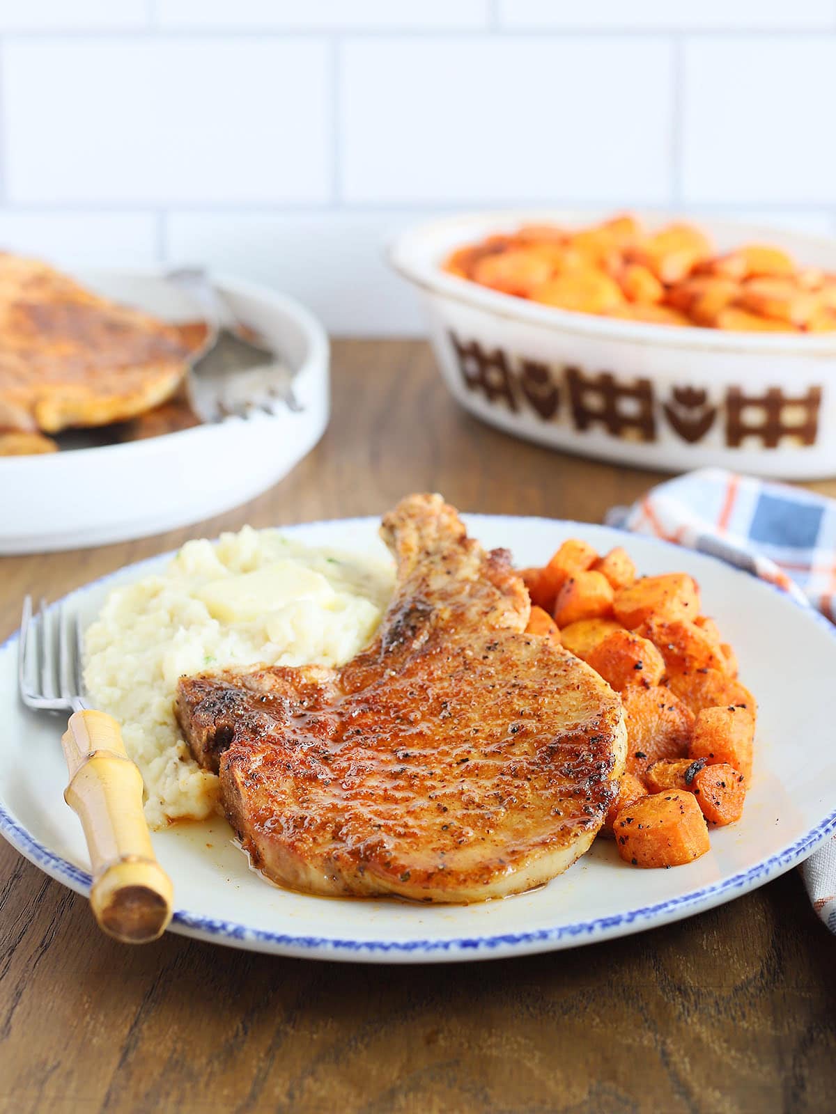 Air fryer bone in pork chop on a plate with mashed potatoes and roasted carrots with a knife and a fork positioned on the side. Serving dishes of carrots and additional pork chops can be seen.