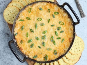 Baked Cajun Catfish Dip garnished with sliced green onions on a wooden cutting board with round sesame seed crackers on the side.