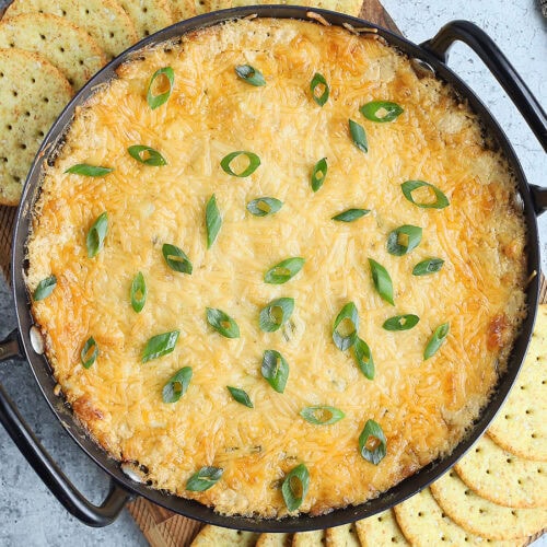 Baked Cajun Catfish Dip garnished with sliced green onions on a wooden cutting board with round sesame seed crackers on the side.