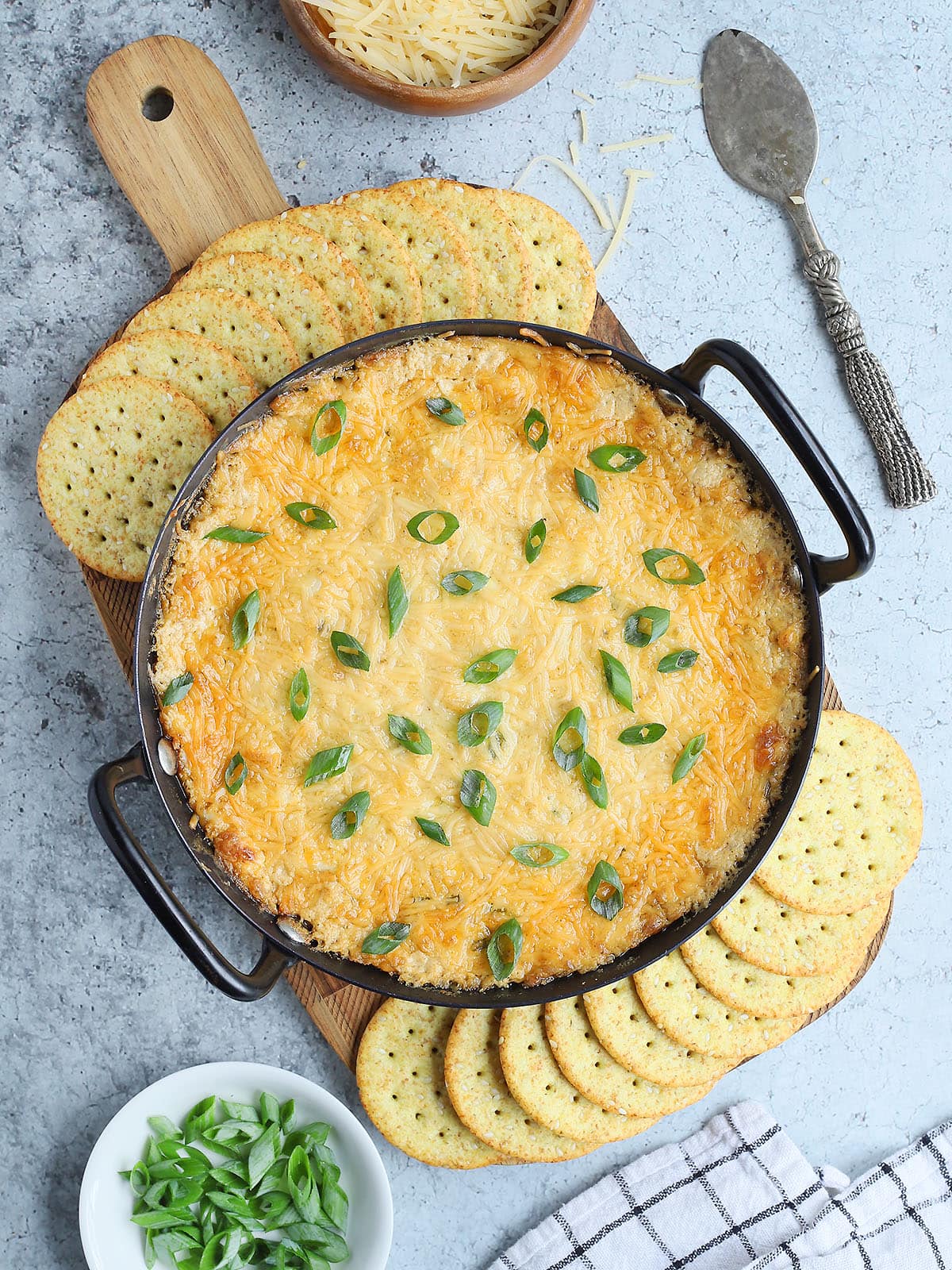 Baked Cajun Catfish Dip garnished with sliced green onions on a wooden cutting board with round sesame seed crackers arranged around the dish.