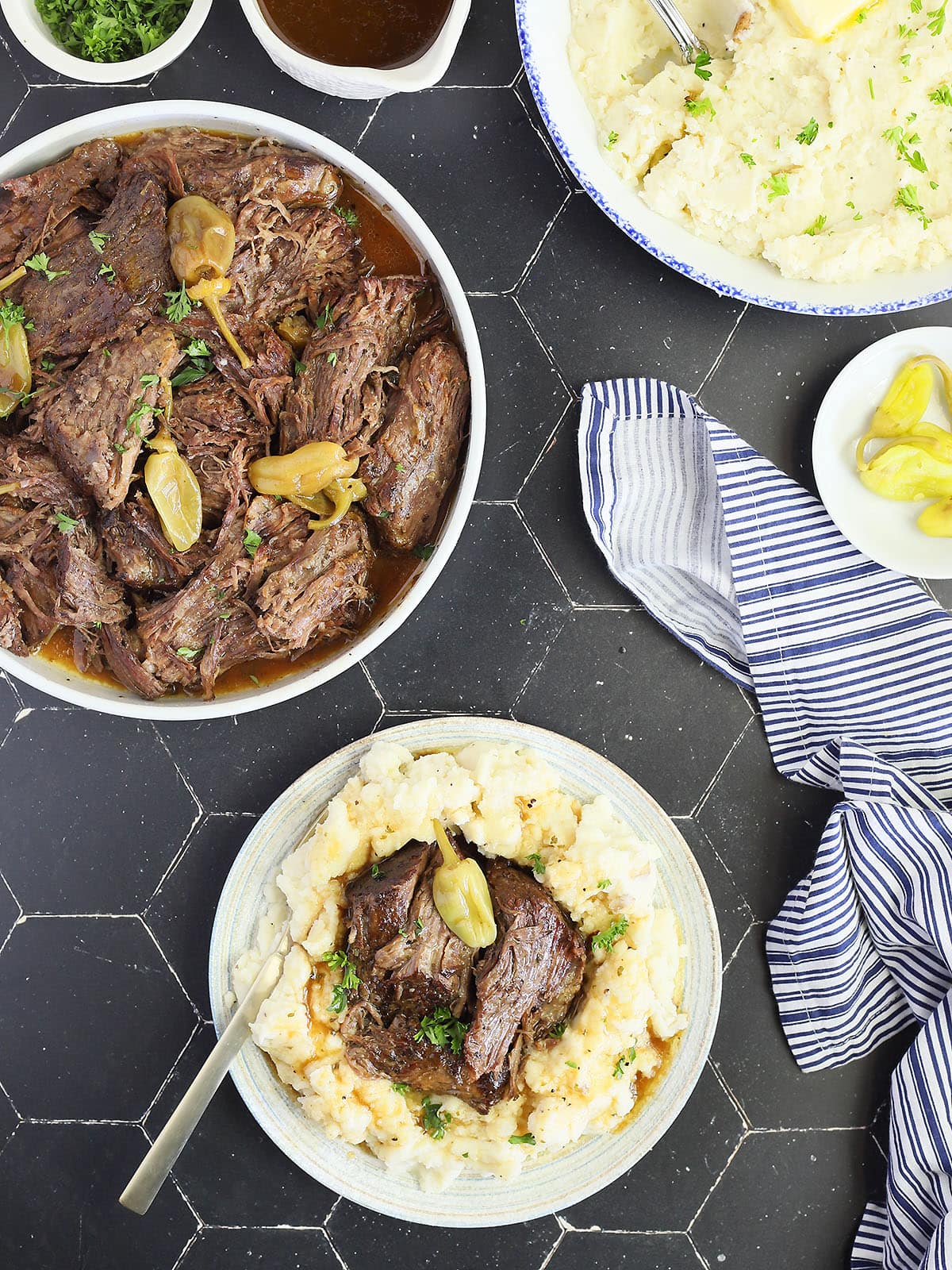 Instant Pot Mississippi Pot Roast served over a bed of mashed potatoes and gravy. A gravy boat, serving dish with roast, a small dish of peppers and an Instant Pot are seen in the background.
