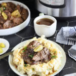 Instant Pot Mississippi Pot Roast served over a bed of mashed potatoes and gravy. A gravy boat, serving dish with roast, a small dish of peppers and an Instant Pot are seen in the background.