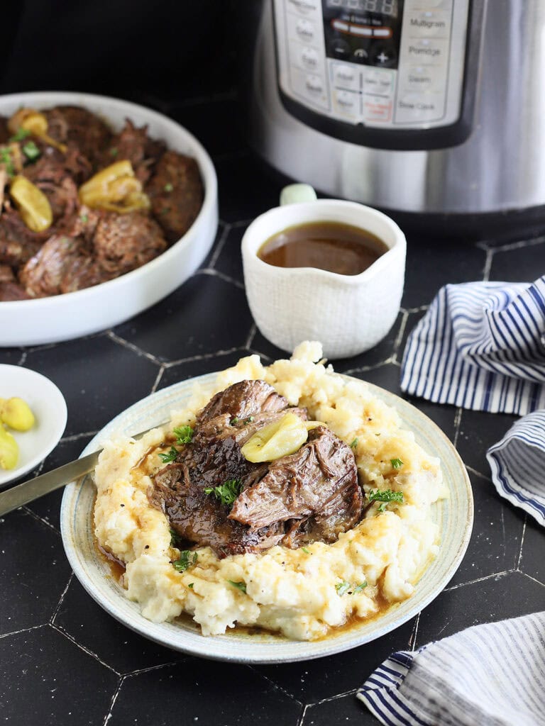 Instant Pot Mississippi Pot Roast served over a bed of mashed potatoes and gravy. A gravy boat, serving dish with roast, a small dish of peppers and an Instant Pot are seen in the background.