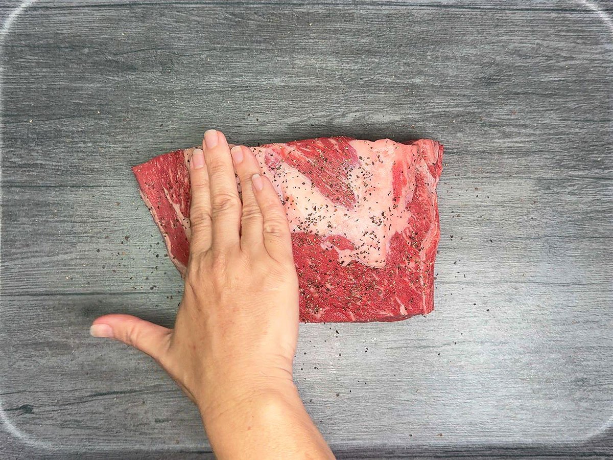 Patting salt and pepper into the surface of a chuck roast.