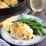 Stuffed catfish fillet on a floral plate with green beans and a lemon wedge. Glass of white wine and a skillet with additional catfish fillets in the background.